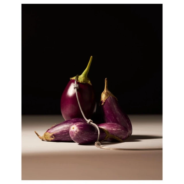 Studio shot of several eggplants on a table, with a silver chain draped across the vegetables against a dark brown background.