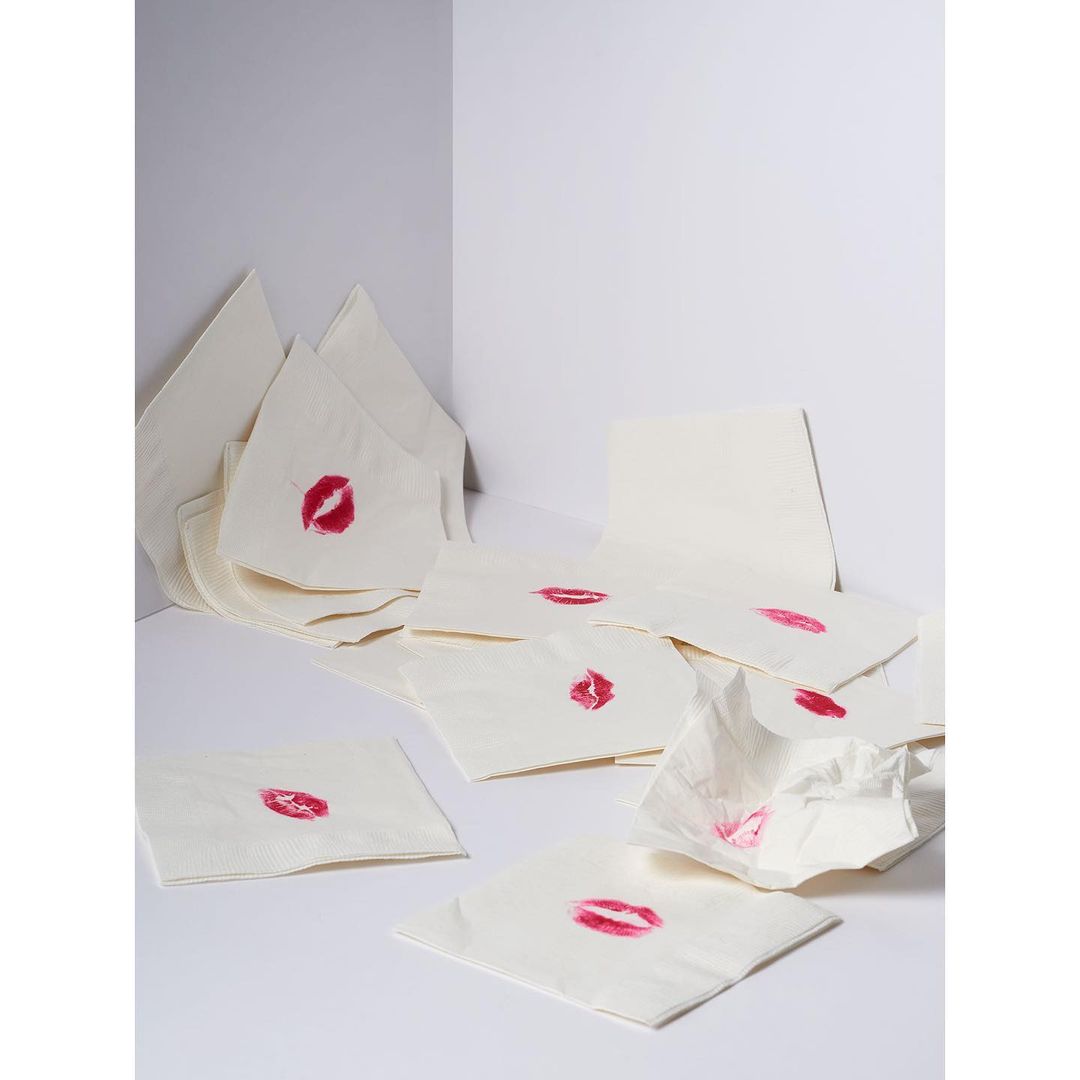 Close-up shot of multiple white napkins scattered around, each bearing a bright red lipstick kiss mark in a clean, white corner setting.