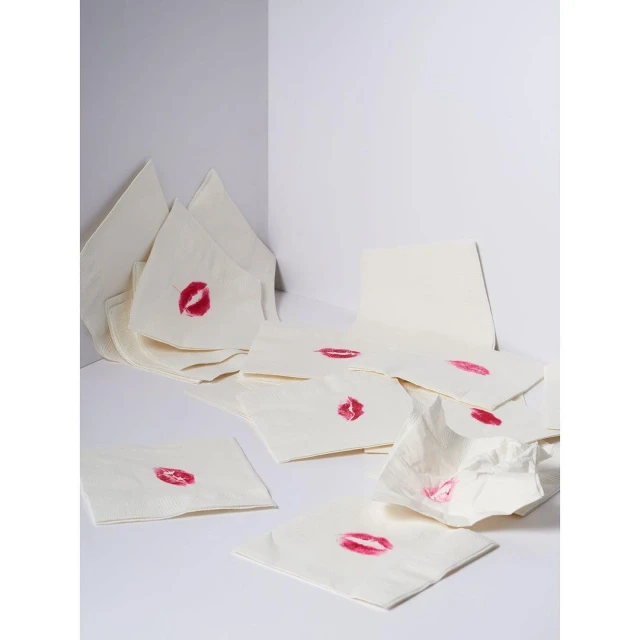 Close-up shot of multiple white napkins scattered around, each bearing a bright red lipstick kiss mark in a clean, white corner setting.
