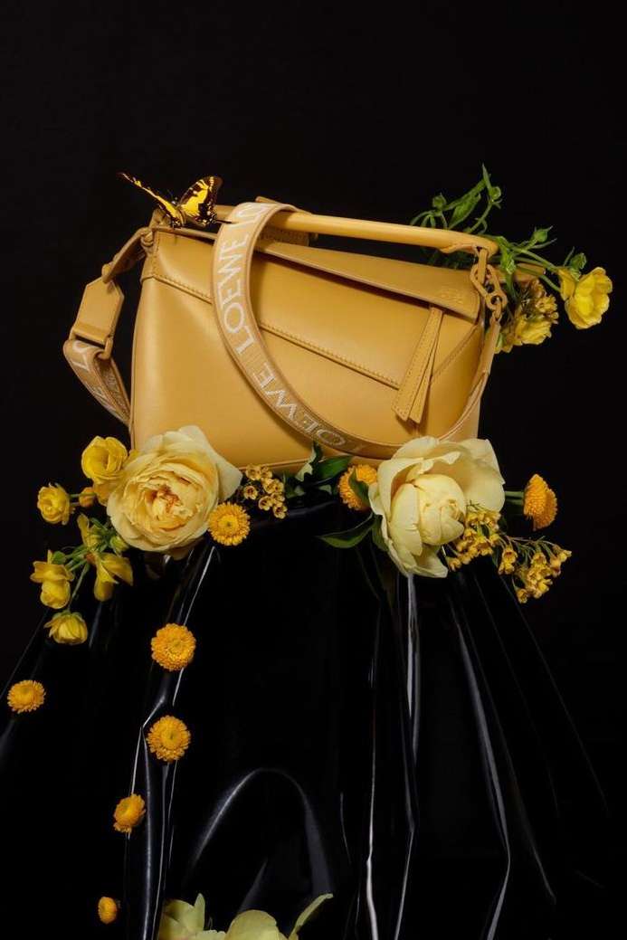 Loewe tan leather handbag displayed with yellow roses and a butterfly against a black background in a studio product shot.