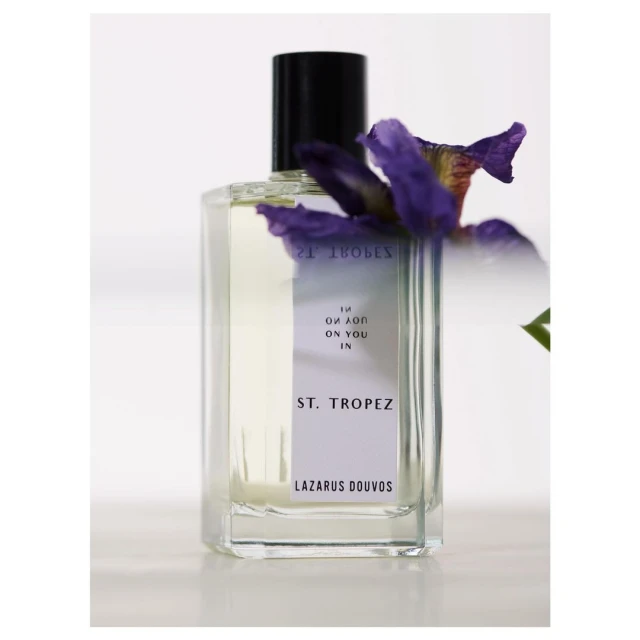 Elegant square perfume bottle with black cap and violet flower accent, minimalist beauty shot against a bright white background.