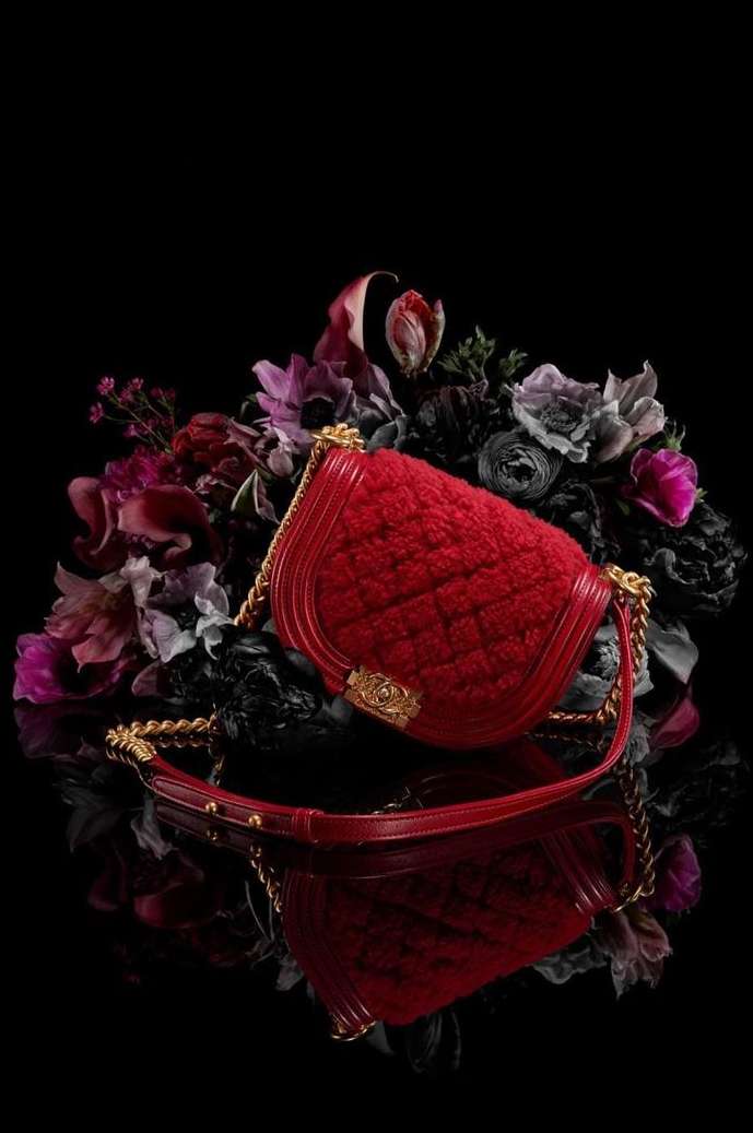 A red Chanel handbag with gold chain strap is displayed on a reflective surface among dark roses and flowers on a dark background.