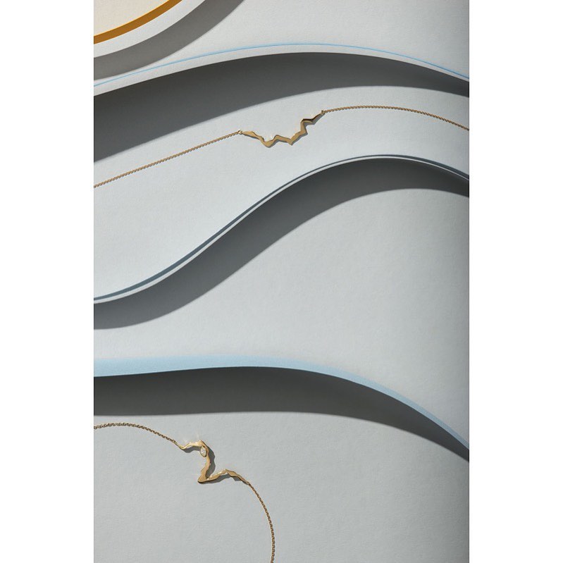Minimalist art featuring abstract gold necklaces against a muted grey and white background with curving lines and shadows.