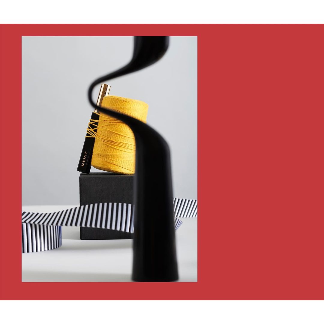 Cosmetic product arrangement featuring a perfume bottle, yellow yarn, ribbon, and black block on a white surface with a red background.