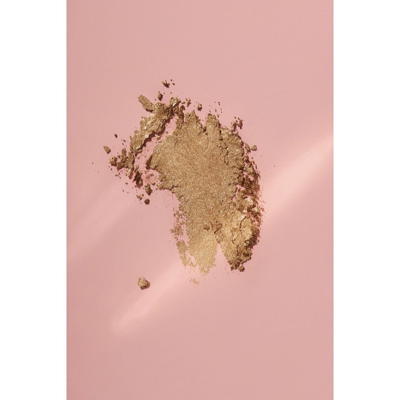 Close-up of golden eye shadow powder on a pale pink background, with soft lighting and subtle gradient adding depth to the scene.