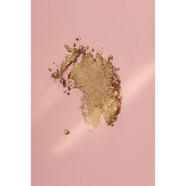 Close-up of golden eye shadow powder on a pale pink background, with soft lighting and subtle gradient adding depth to the scene.