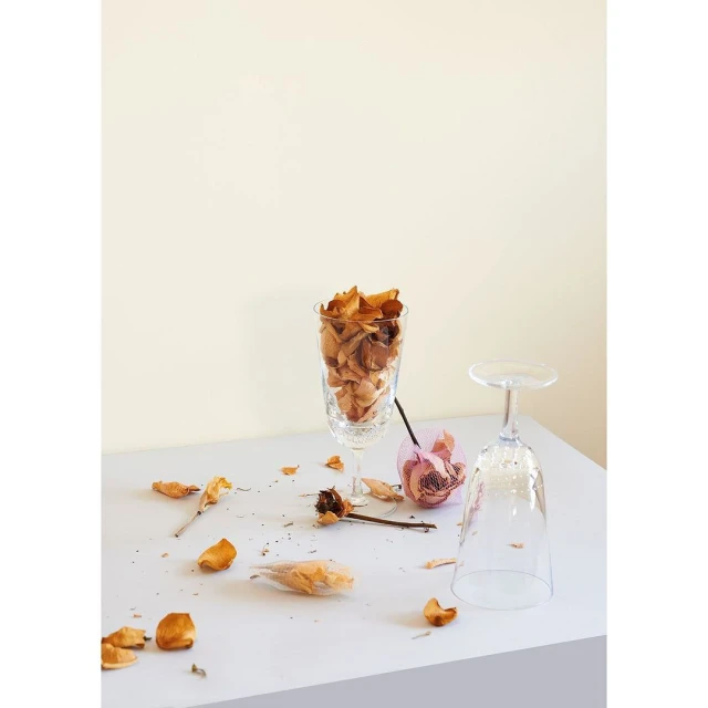 Still life with dried rose petals and clear decorative glasses on a white surface against a plain cream-colored background.
