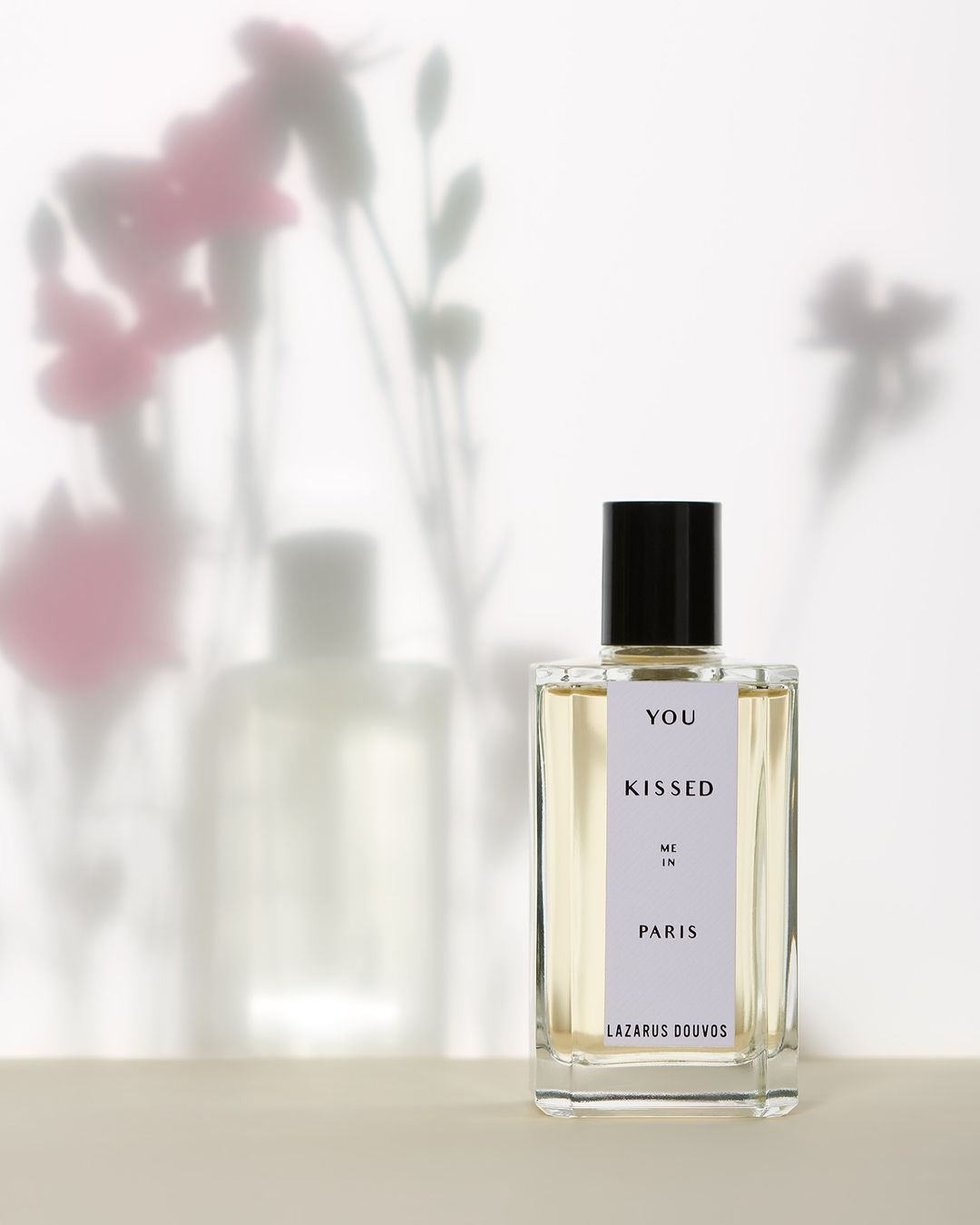 Product shot of a 'You Kissed Me In Paris' perfume bottle by Lazarus Douvos on a beige surface with flower shadows on the white backdrop.