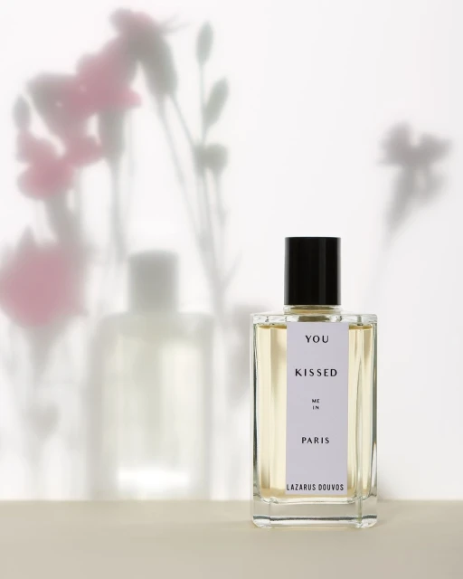 Product shot of a 'You Kissed Me In Paris' perfume bottle by Lazarus Douvos on a beige surface with flower shadows on the white backdrop.