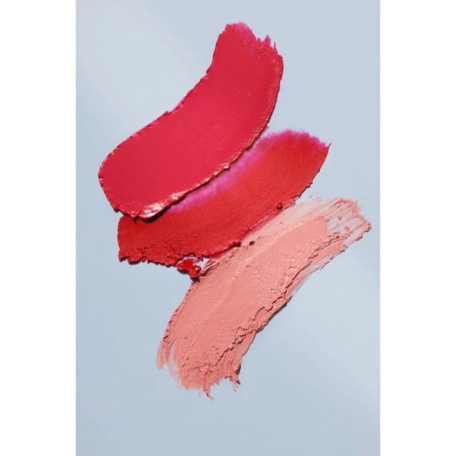 Three swatches of lipstick in red, coral, and pink colors are arranged diagonally against a light blue background, showcasing texture.
