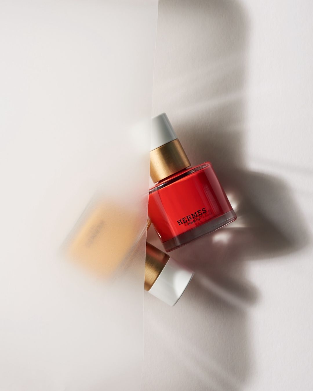 Close-up of a red Hermès nail polish bottle with gold accents against a bright white background, soft lighting and shadow details.