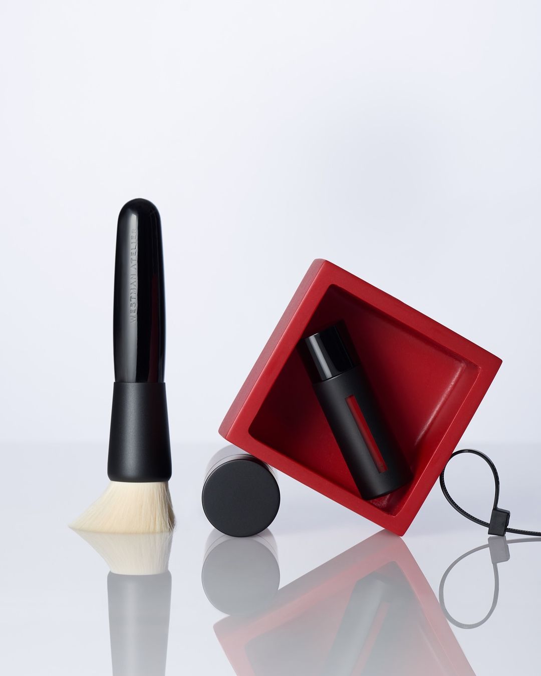 A makeup brush, black jar, and black bottle are arranged next to a red square box on a reflective white surface against a white background.