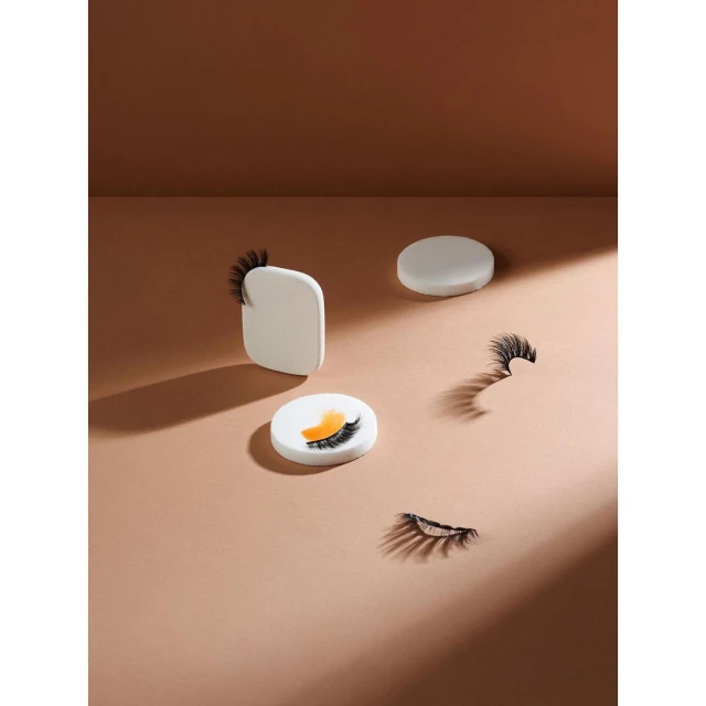 Arrangement of individual false eyelashes on small pedestals against a neutral backdrop, lit to create shadows and highlight the products.