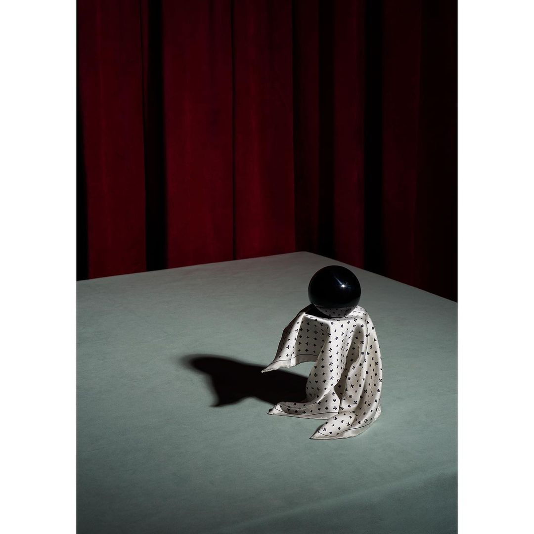 Still life of a black sphere on a white polka dot cloth, draped on a table against a red curtain backdrop, lit by studio lighting.