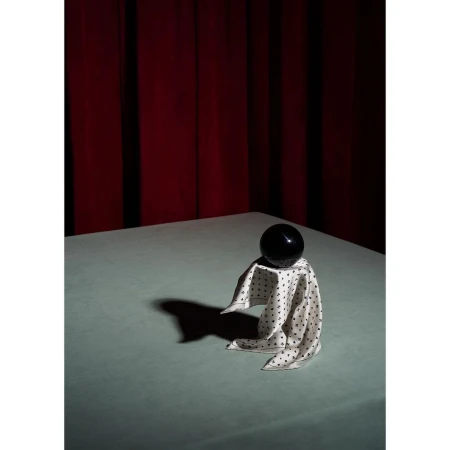 Still life of a black sphere on a white polka dot cloth, draped on a table against a red curtain backdrop, lit by studio lighting.