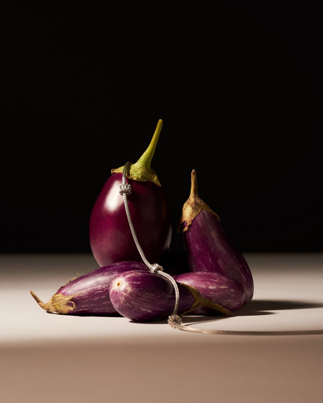 Still life studio shot of purple eggplants arranged on a beige surface against a dark background, with a silver chain.