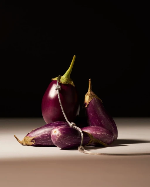 Still life studio shot of purple eggplants arranged on a beige surface against a dark background, with a silver chain.