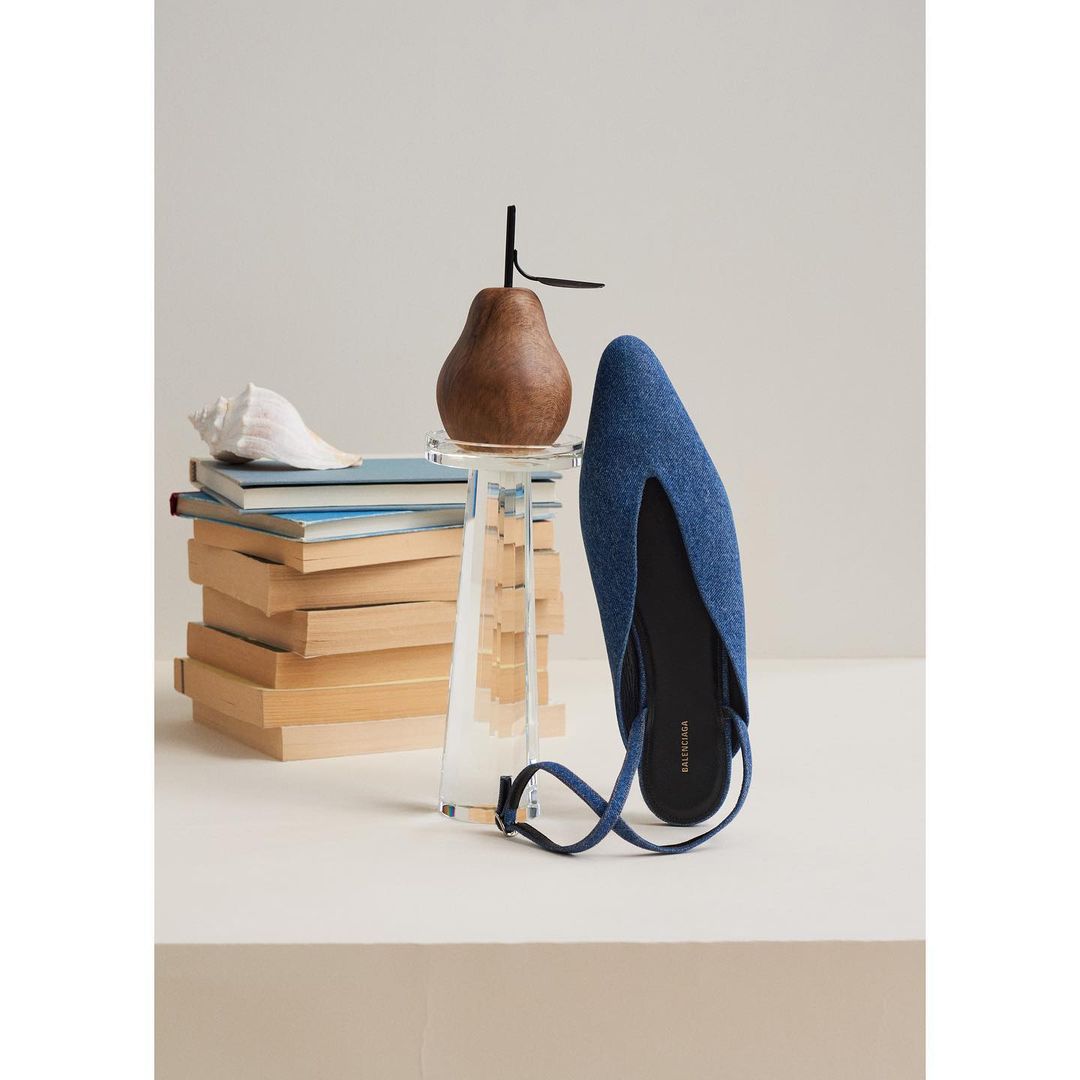Denim-look mule shoe with wood pear on an acrylic stand, books and a seashell against a neutral backdrop in a fashion product shot.
