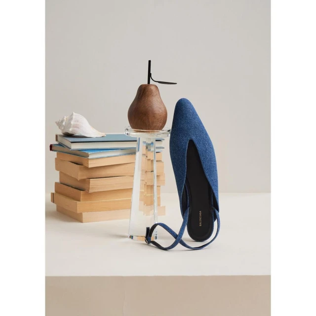 Denim-look mule shoe with wood pear on an acrylic stand, books and a seashell against a neutral backdrop in a fashion product shot.