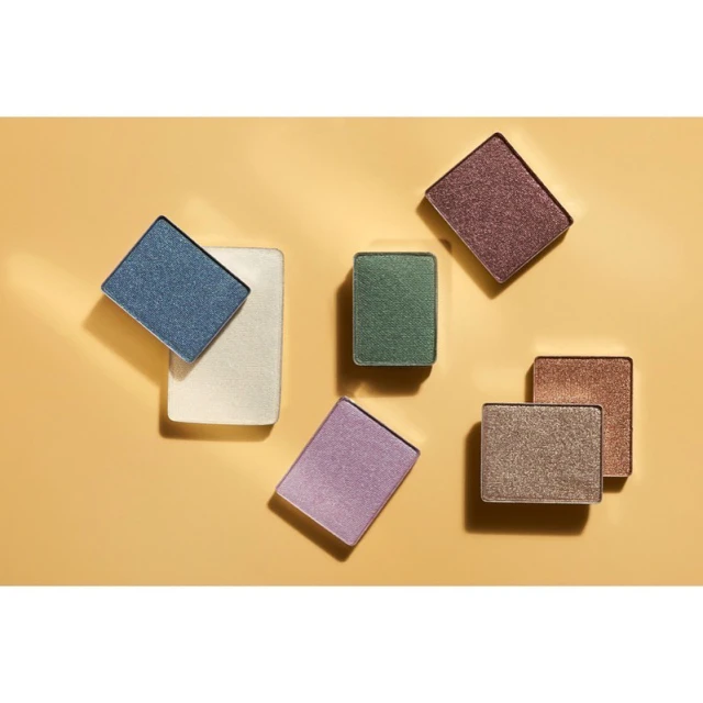 Overhead shot of arranged eyeshadow singles in various colors, including blue, green, purple, and brown, on a tan background.