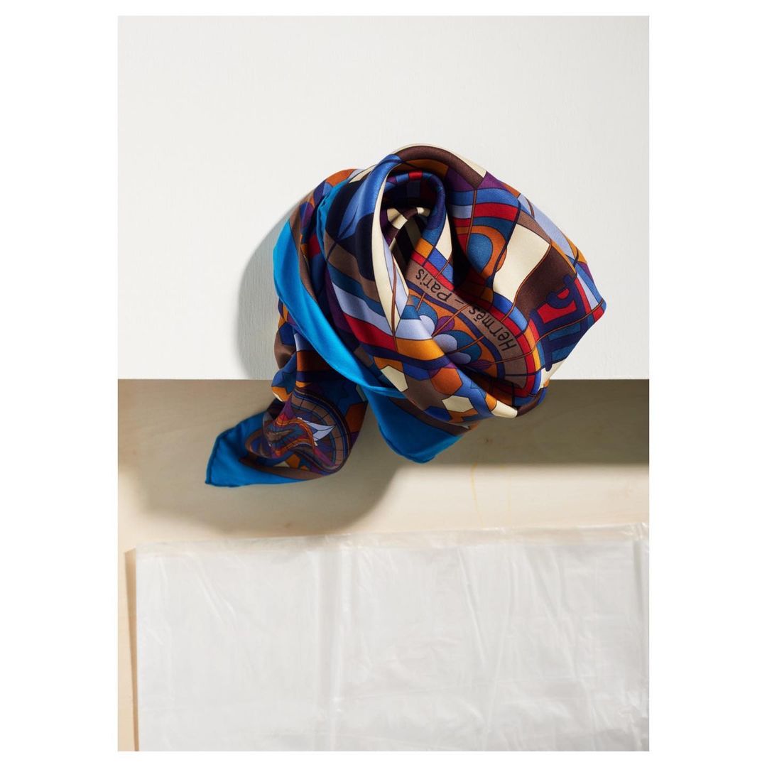 Stylized Hermès silk scarf with vibrant blue, red, and beige patterns, draped on a white surface, displaying luxury fashion.