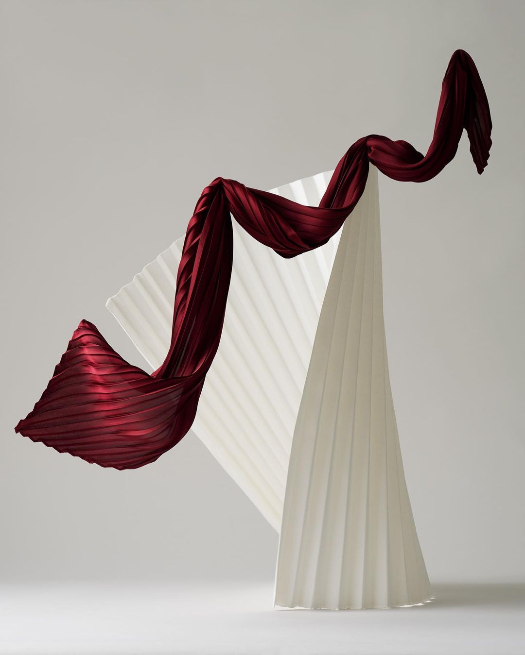 Abstract product shot with a burgundy pleated fabric draped over a white, pleated geometric pedestal against a gray gradient background.