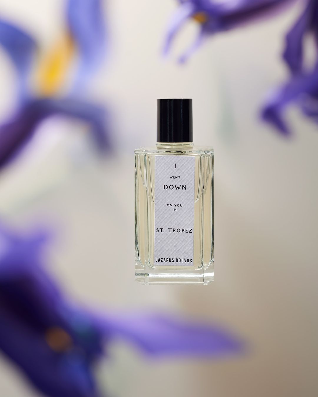 Rectangular perfume bottle with a black cap, partially framed by blurred purple flowers, against a neutral light-grey background.