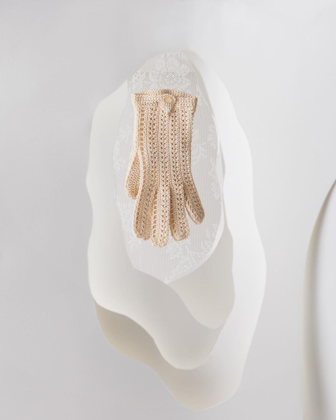 Close-up of a single, cream-colored, openwork crochet glove on a draped, mannequin-like form against a neutral white backdrop.