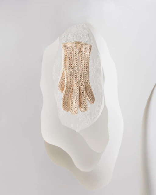 Close-up of a single, cream-colored, openwork crochet glove on a draped, mannequin-like form against a neutral white backdrop.