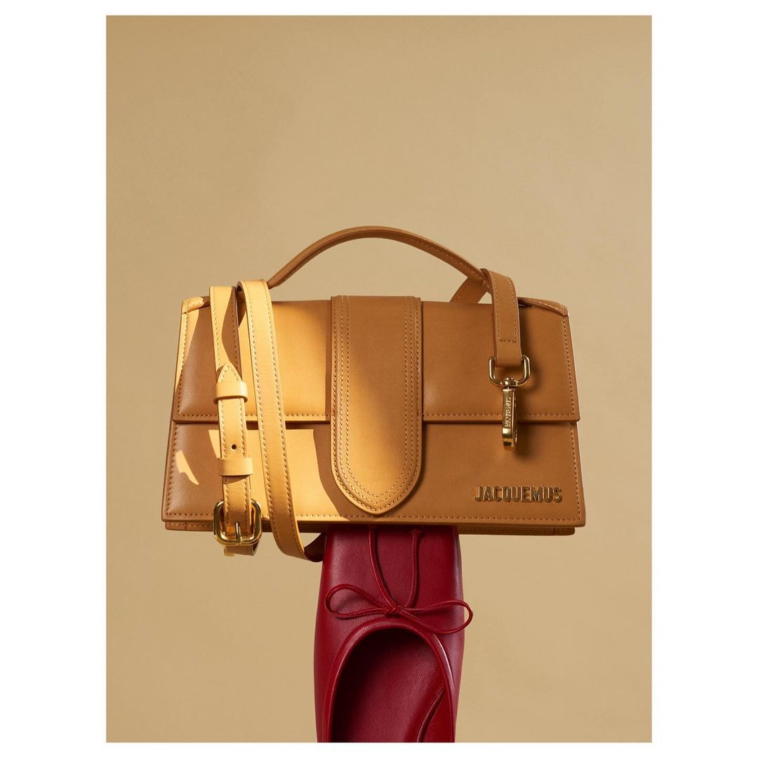 Tan leather Jacquemus handbag with handle and strap, balanced on a red leather ballet shoe with bow against a neutral background.