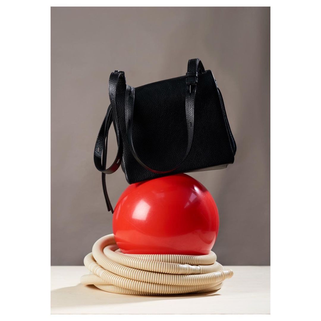 Studio product shot of a black purse balanced on top of a large red ball and a beige coiled cable on a white surface.