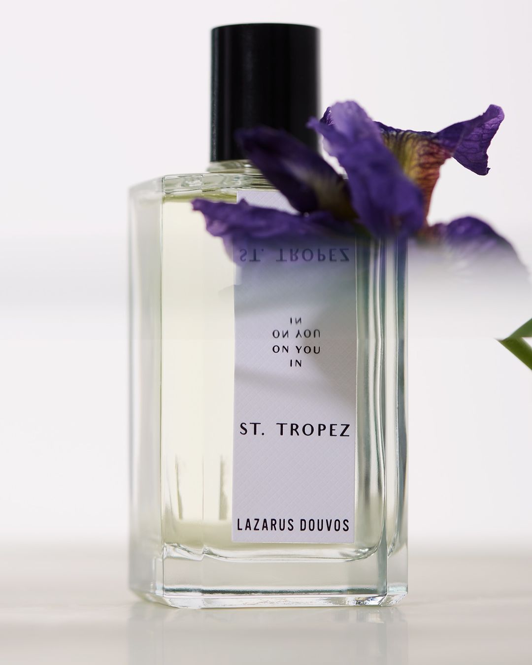Perfume bottle of St. Tropez Lazarus Douvos with a clear bottle, black cap, and purple flower against a white background.