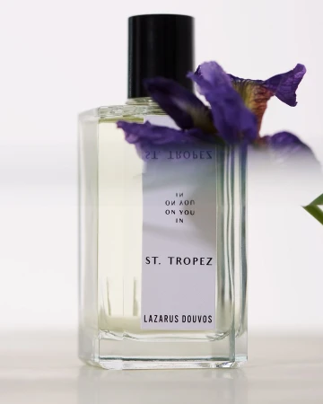 A rectangular glass perfume bottle with a black cap and white label rests against a clean white background with a purple flower