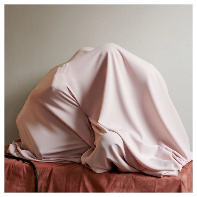 Abstract studio shot of a person covered by a light pink fabric on top of a table against a light-colored wall.