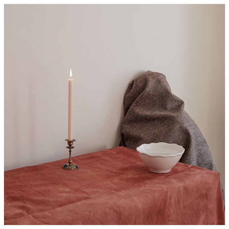 Still life featuring a lit pink candle in a brass holder on a coral tablecloth next to a gray drape and white bowl indoors.
