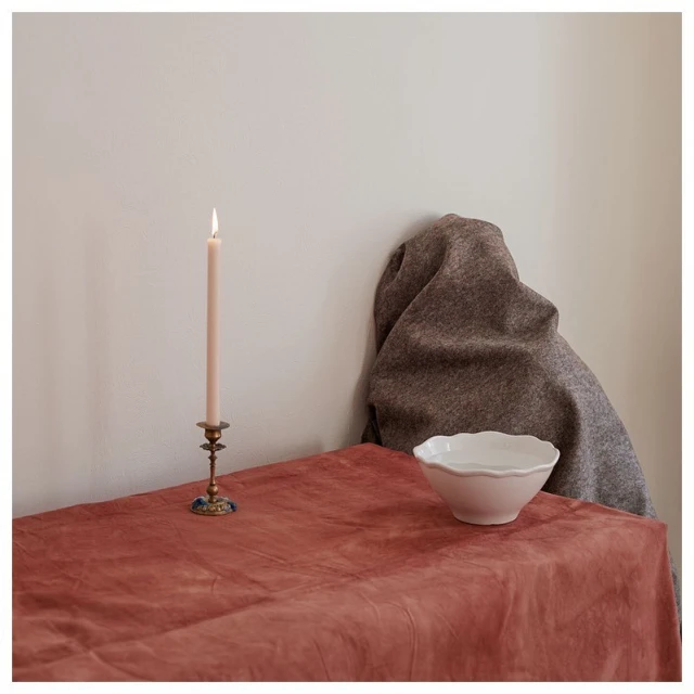 Still life featuring a lit pink candle in a brass holder on a coral tablecloth next to a gray drape and white bowl indoors.