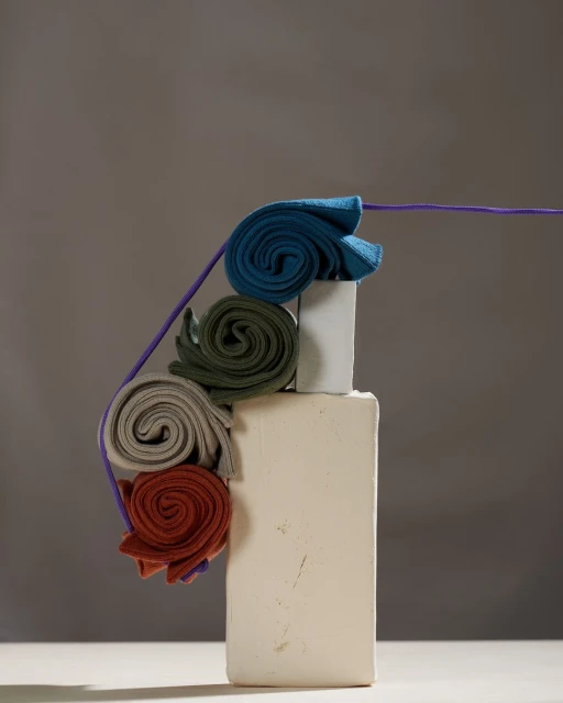 Still life of rolled textiles on white block with a purple cord, creating a textured and layered effect on neutral background.