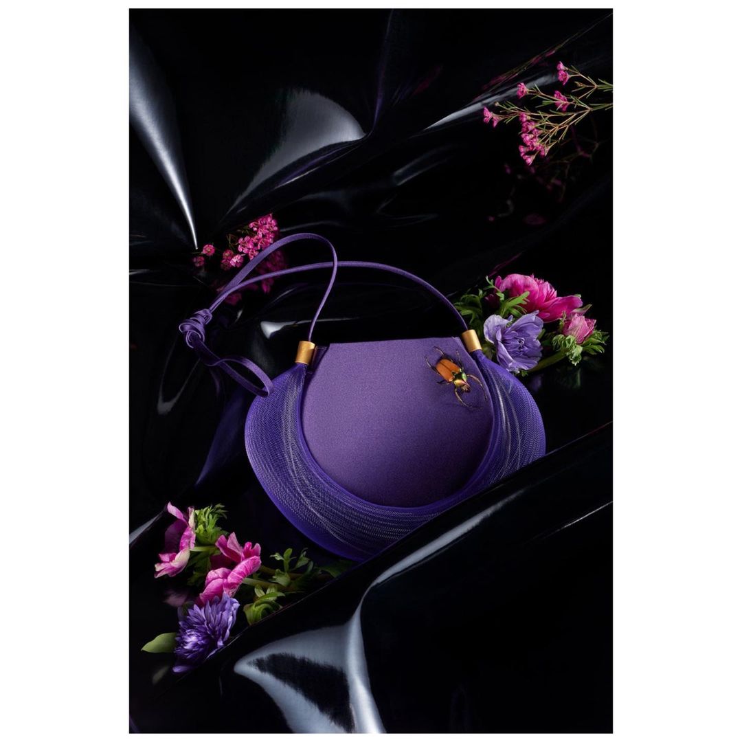 Elegant purple handbag with tassel detail and an ornamental bug brooch, styled with pink and purple floral arrangements against a black glossy backdrop.