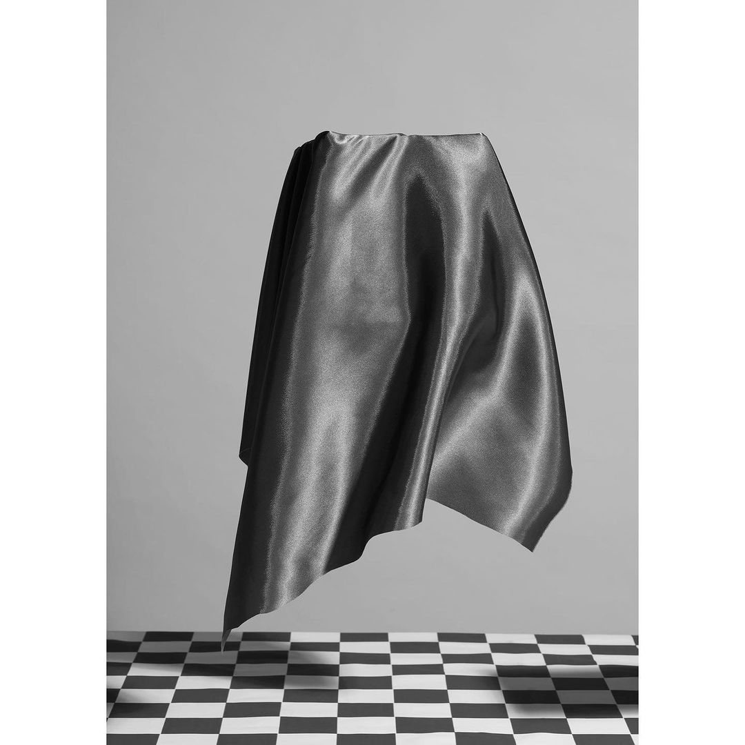 Draped gray satin fabric cascades over an unseen object, floating above a black and white checkered floor against a light gray backdrop.