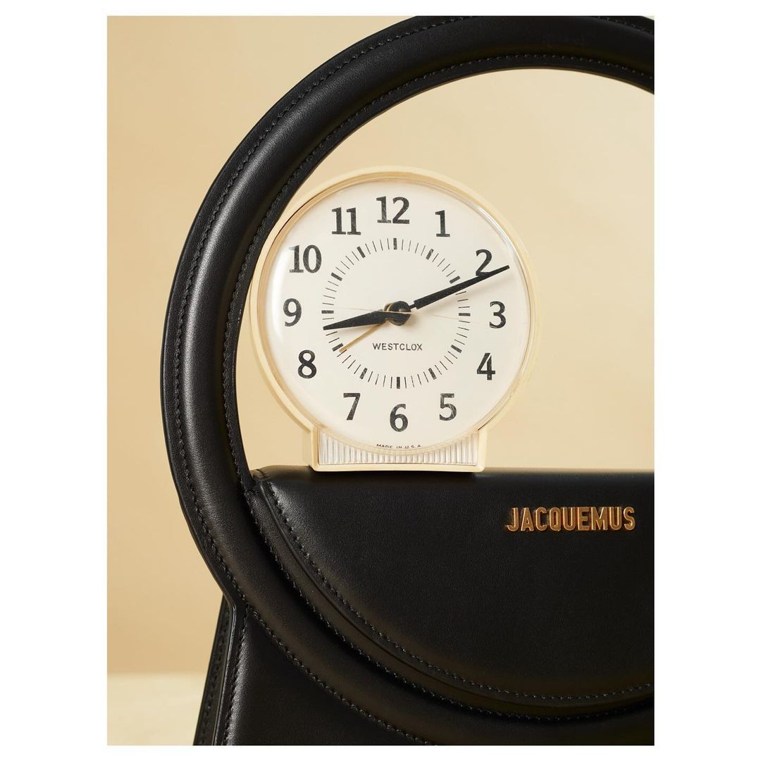 Close-up studio shot of a black leather handbag with a round top handle, featuring a small, round clock placed on top of the bag.