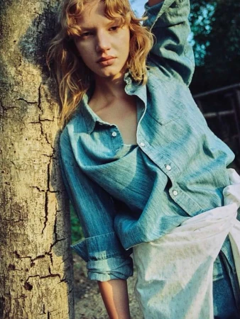 A young woman with blonde curly hair wearing a blue denim shirt leaning against a tree trunk in an outdoor setting.