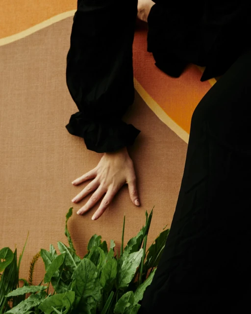 Close-up of a hand gently resting above green leaves and sprouts against a textured tan and orange backdrop with black clothing accents.