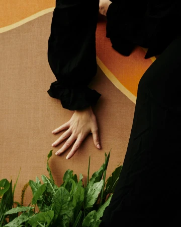 Close-up of a hand gently resting above green leaves and sprouts against a textured tan and orange backdrop with black clothing accents.
