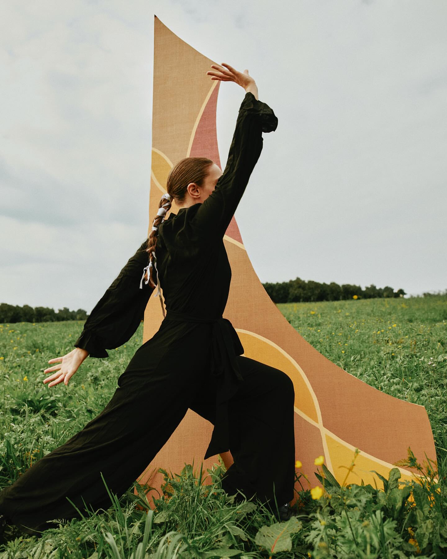 Adult woman in a black jumpsuit poses in a field of tall grass beside a large patterned backdrop under a cloudy sky.