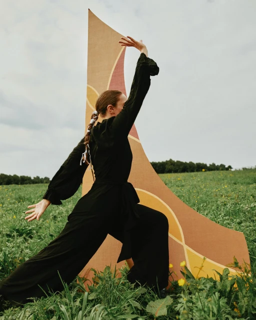 Adult woman in a black jumpsuit poses in a field of tall grass beside a large patterned backdrop under a cloudy sky.