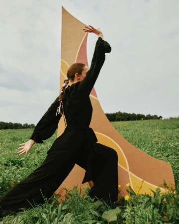 Adult woman in a black jumpsuit poses in a field of tall grass beside a large patterned backdrop under a cloudy sky.