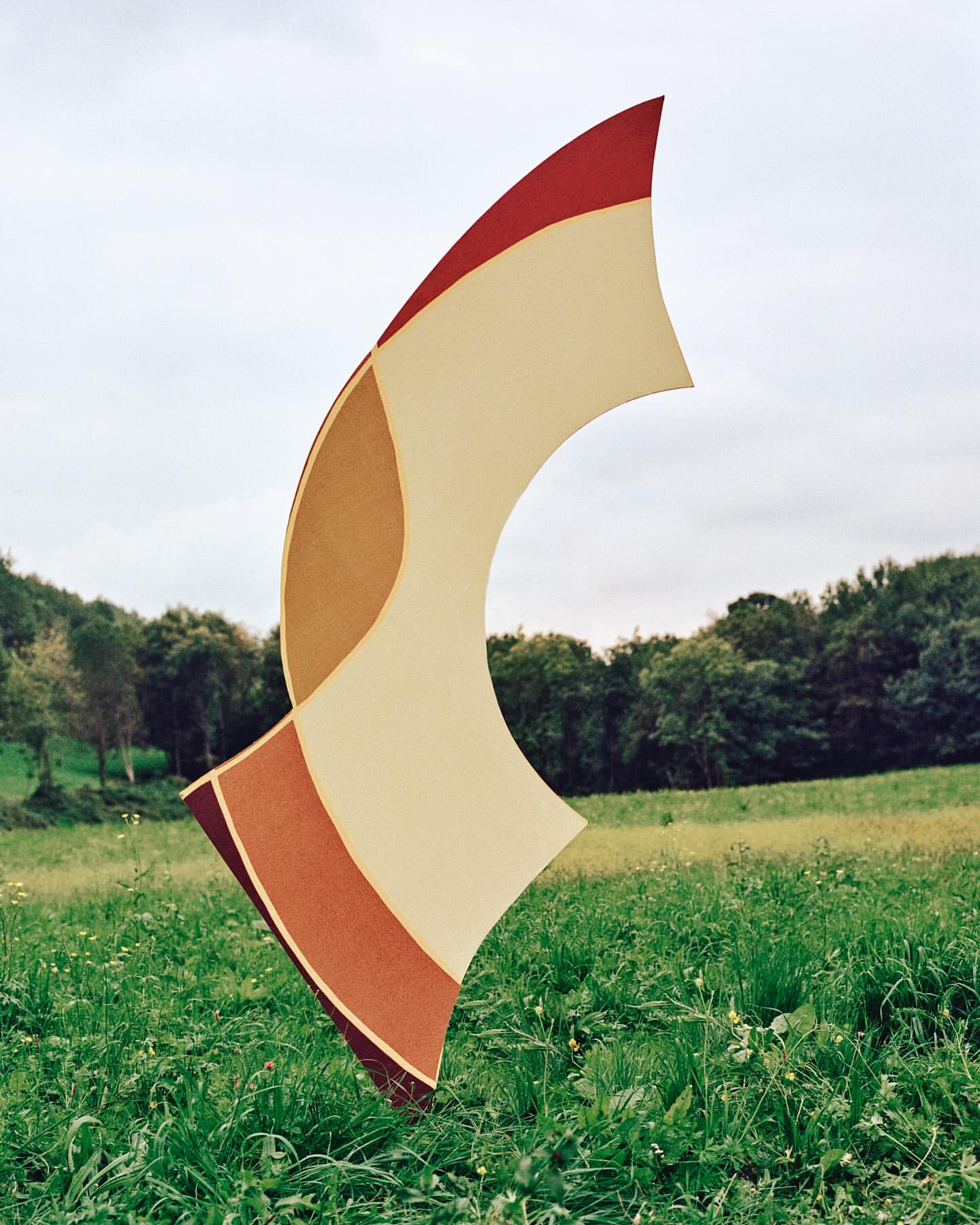 Abstract art sculpture in a green meadow with trees and sky. Red, tan, and brown color bands on the modern art piece.