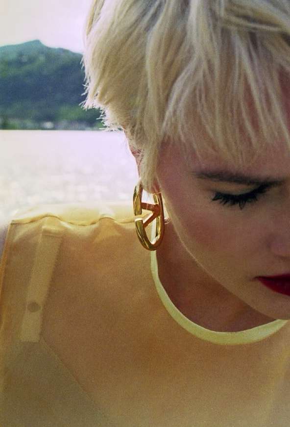 Close up portrait of a blonde woman wearing gold hoop earrings and a translucent yellow top outdoors near water.