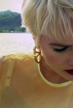 Close up portrait of a blonde woman wearing gold hoop earrings and a translucent yellow top outdoors near water.