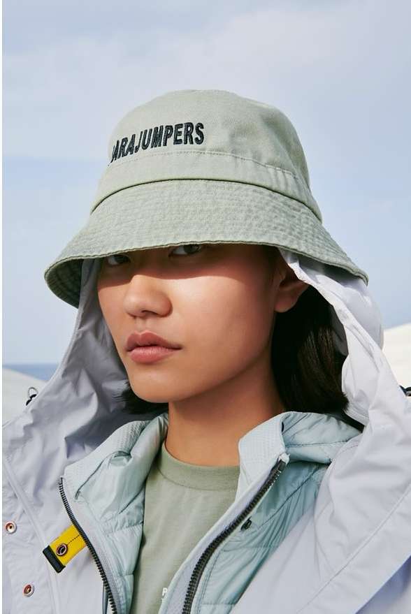 Portrait of a young Asian woman wearing a light-green bucket hat with the 'Parajumpers' logo and a light-blue hooded jacket against an outdoor background.